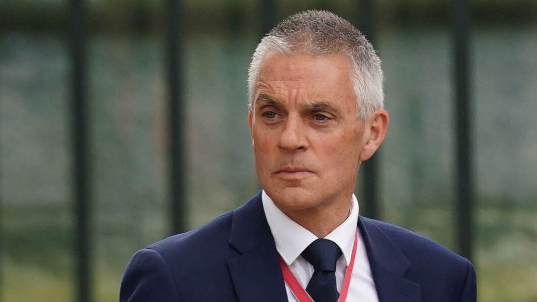 
                    A close crop of Tim Davie shows his head and shoulders as he frowns looking away from camera while wearing a suit and tie and a red lanyard around his neck, pictured outs