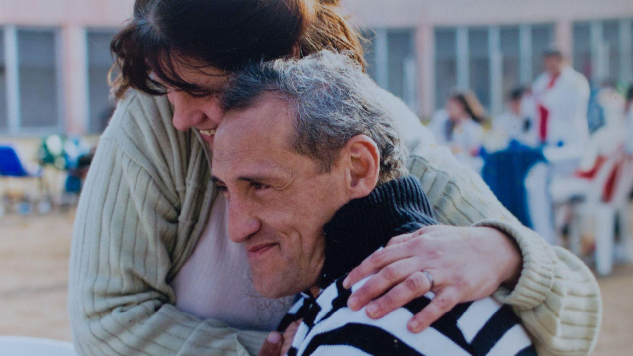 Aleho sedi i izgleda zadovoljno dok ga Andrea, koja stoji, grli sa strane, stavlja levu ruku na njegovo rame i osmehuje se dok naslanja glavu na njegovu.