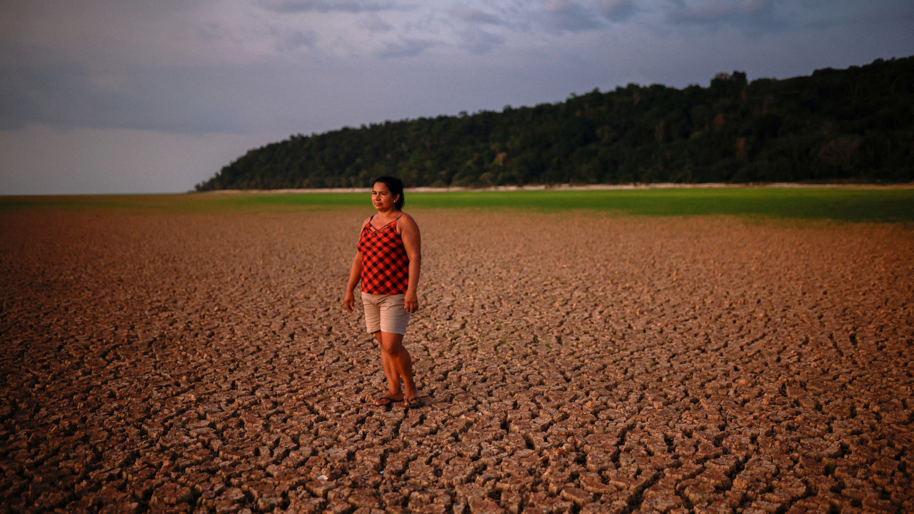 žena stoji na isušenom i ispucalom rečnom koritu dok se u daljini nazire voda 