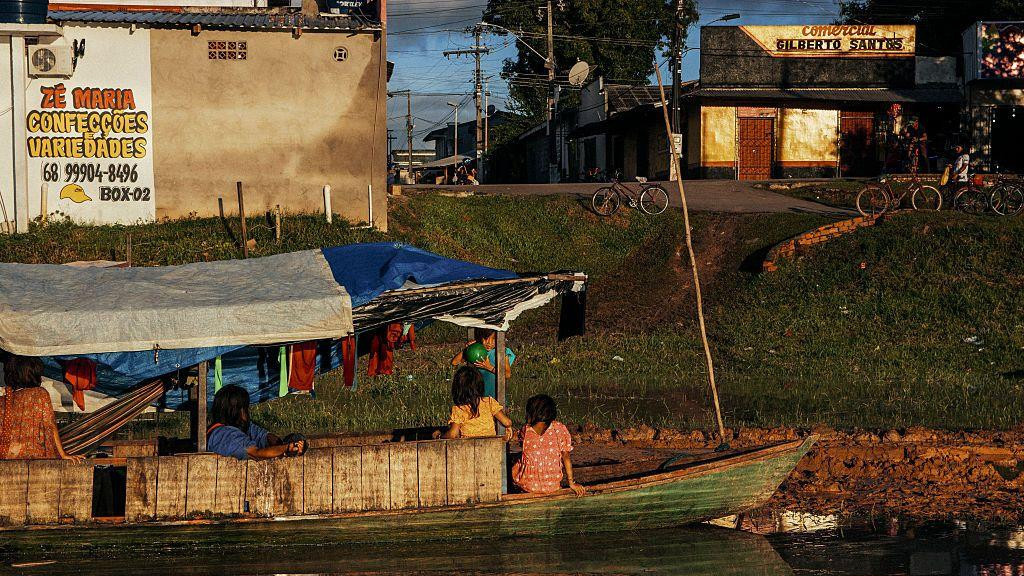 nekoliko ljudi sedi u drvenom čamcu u reci na obali je put i zgrade 