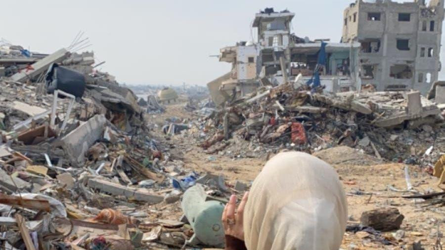 Žena sa maramom na glavi i sivo-smeđem kaputu stoji leđima okrenuta kameri. Ispred nje su ostaci dve uništene betonske zgrade i velike gomile šuta i zemlje