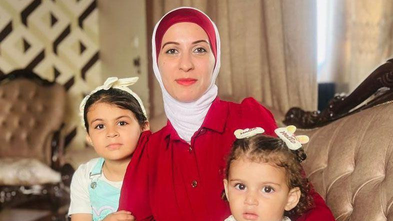 Žena u crvenoj haljini i belo-crvenoj marami na glavi sedi sa dve devojčice