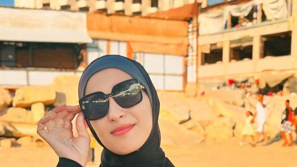 Žena sa velikim naočarama za sunce i crno-sivom maramom na glavi sedi na stolici na plaži. Iza nje se vide uništene zgrade