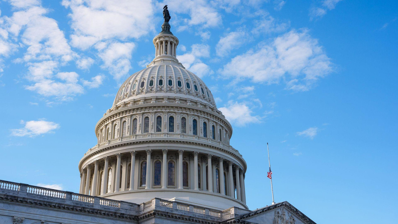 Image shows US Capitol dome against a blue sky with partial cloud cover and the US flag flying in the foreground. Photo taken in Washington DC on Monday.