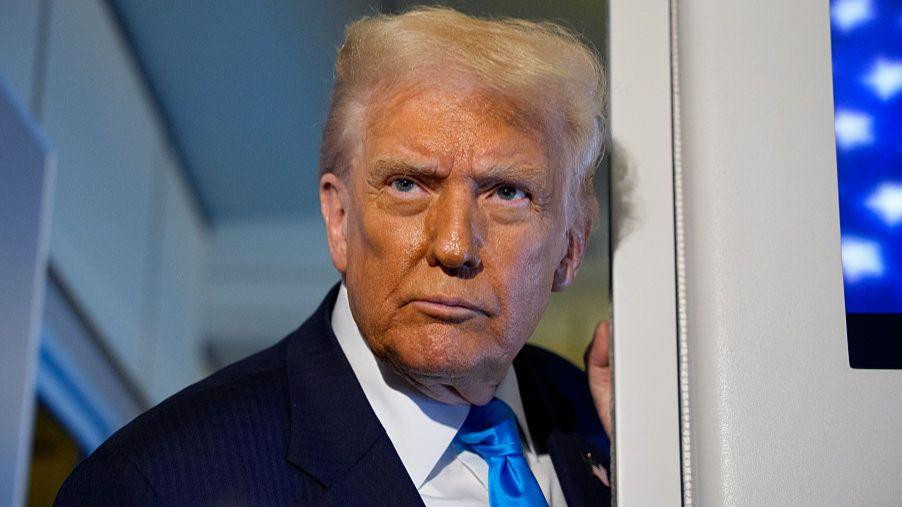 
                    US president donald trump is framed in a doorway on Air Force One as he speaks to reporters. It's a close up shot, revealing what looks like fake tan on his face. he is w