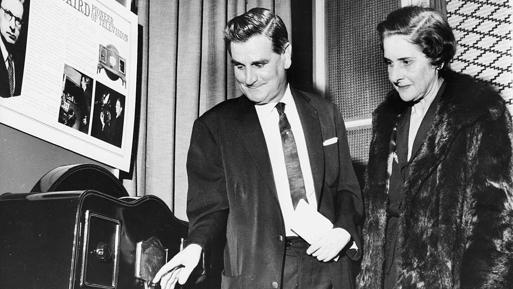 Vilijem Tejton u odelu pokazuje rukom na radio Margaret Beird, koja je u bundi i gleda