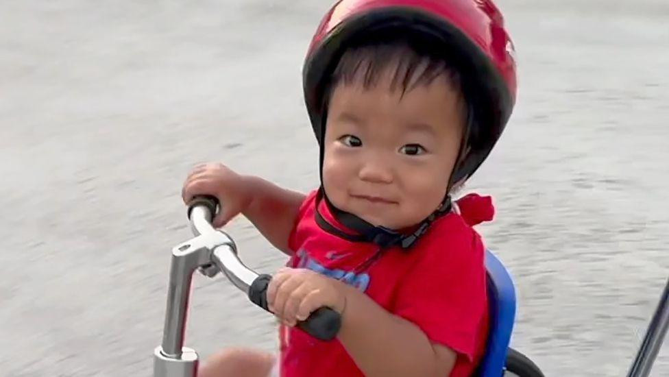 Oliver Chu rides a red tricycle. He is wearing a red helmet, red T-shirt and is looking at the camera