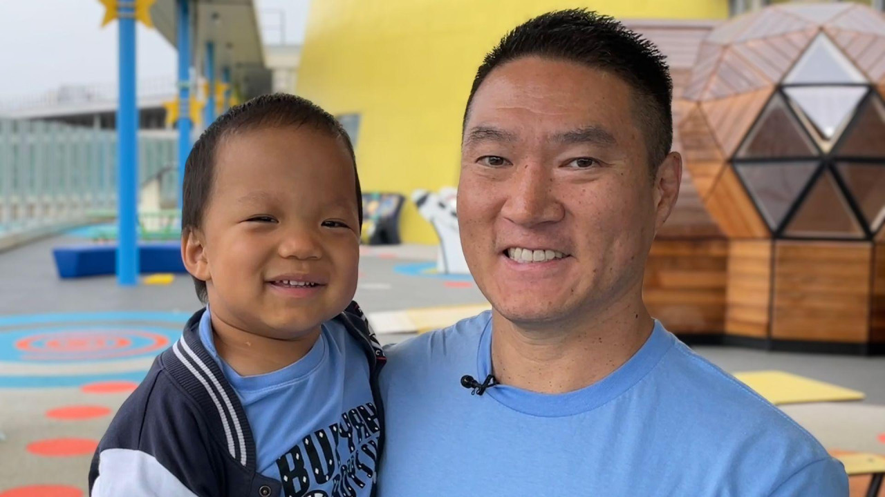 Tata Riki nosi Olivera u naručju. Smeju se i nose plave majice. Nalaze se na krovu sa šarenim sedištima i stolovima u pozadini.