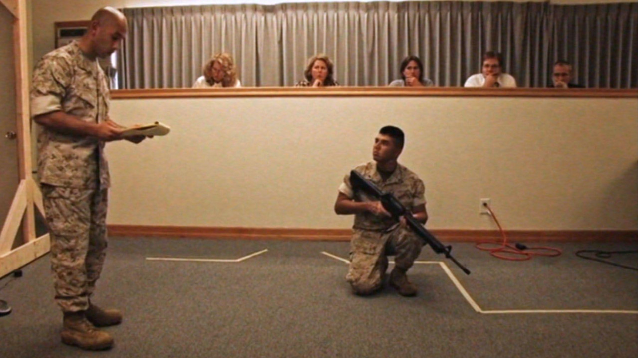 s desne strane muškarac u vojnoj opremi i sa puškom u ruci kleči. Gleda u muškarca spram sebe koji takođe nosi vojnu odeću. Iza njih sedi petoro ljudi jedni pored drugih 