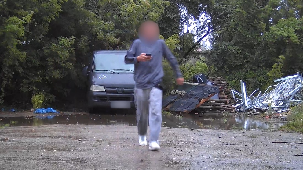 
                    A surveillance photo of a male teenager wearing grey sweat pants and shirt and with his face blurred, carrying a mobile phone. He is walking away from a grey van parked o
