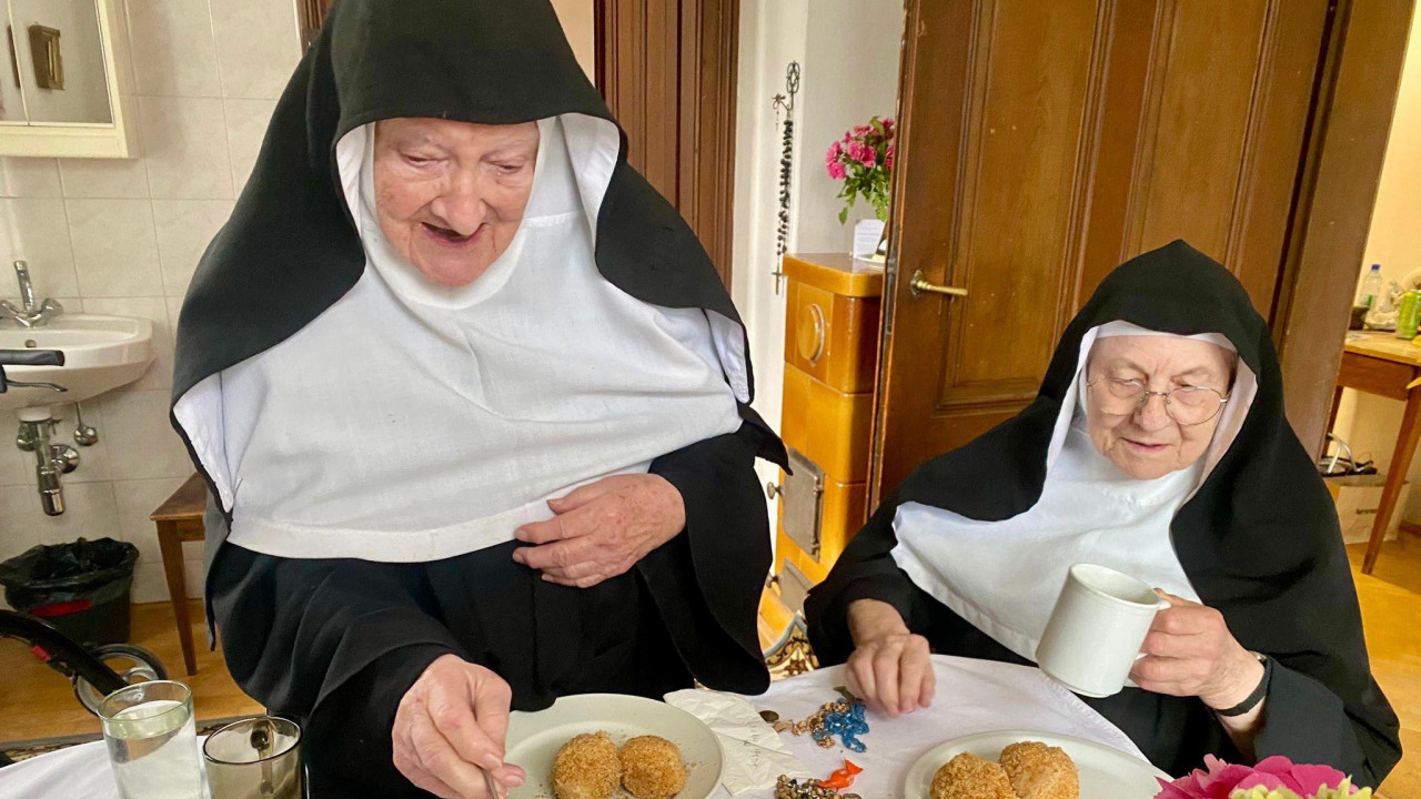 Dve časne sestre jedu, jedna služi drugu hranom, časne sestre jedu knedle sa šljivama, deve starije žene obučene kao časne sestre jedu knedle sa šljivama 