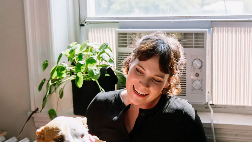Devojka tamnije vezane kose sedi pored prozora i u krilu drži psa, iza nje su zelena biljka i radijator