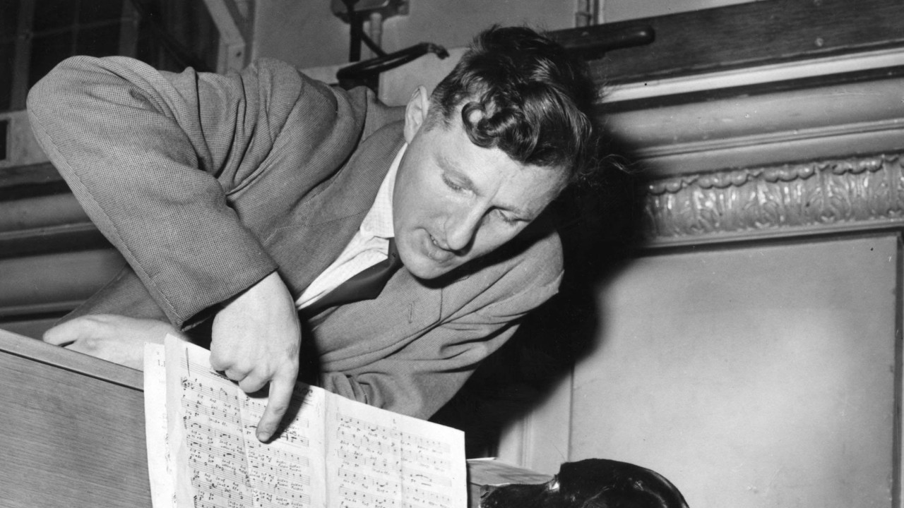 Pas sedi za klavirom i svira, gleda u note dok mu muškarac prstom pokazuje u kajdanku, crnobela fotografija