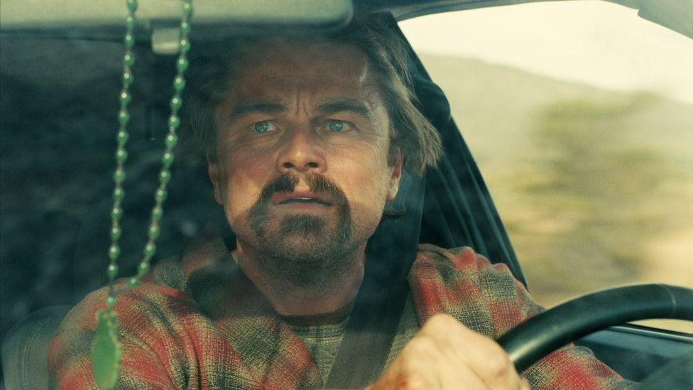 Leonardo Di Kaprio u filmu „Jedna bitka za drugom“, vozi auto i deluje zabrinuto dok gleda u retrovizor, scena iz filma One Battle After Another