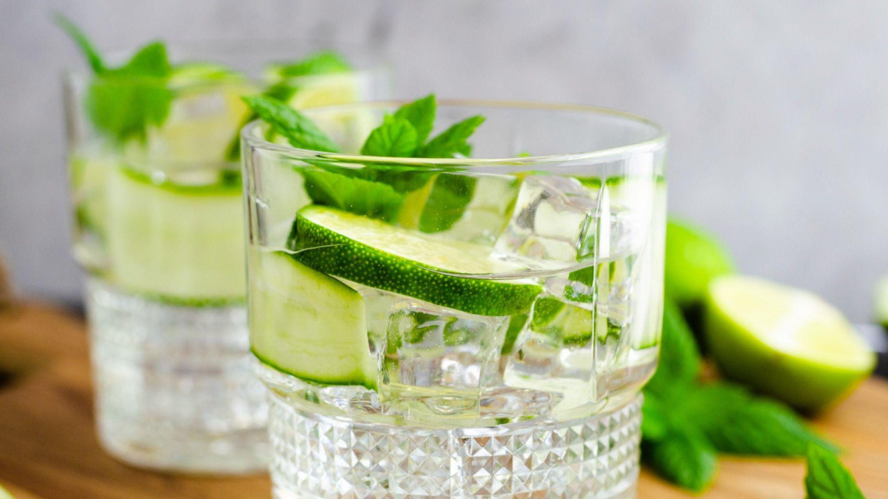 Two glasses of gin and tonic with sliced lime and mint, resting on a wooden board. Sliced lime and mint leaves are lying next to the glasses.