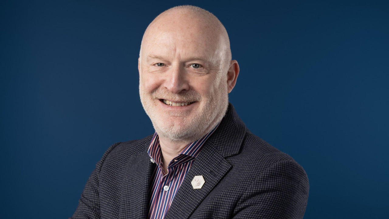 Man with white stubble on his chin smiles at the camera. He's wearing a shirt and tweed jacket. The background is a blue photographic screen 