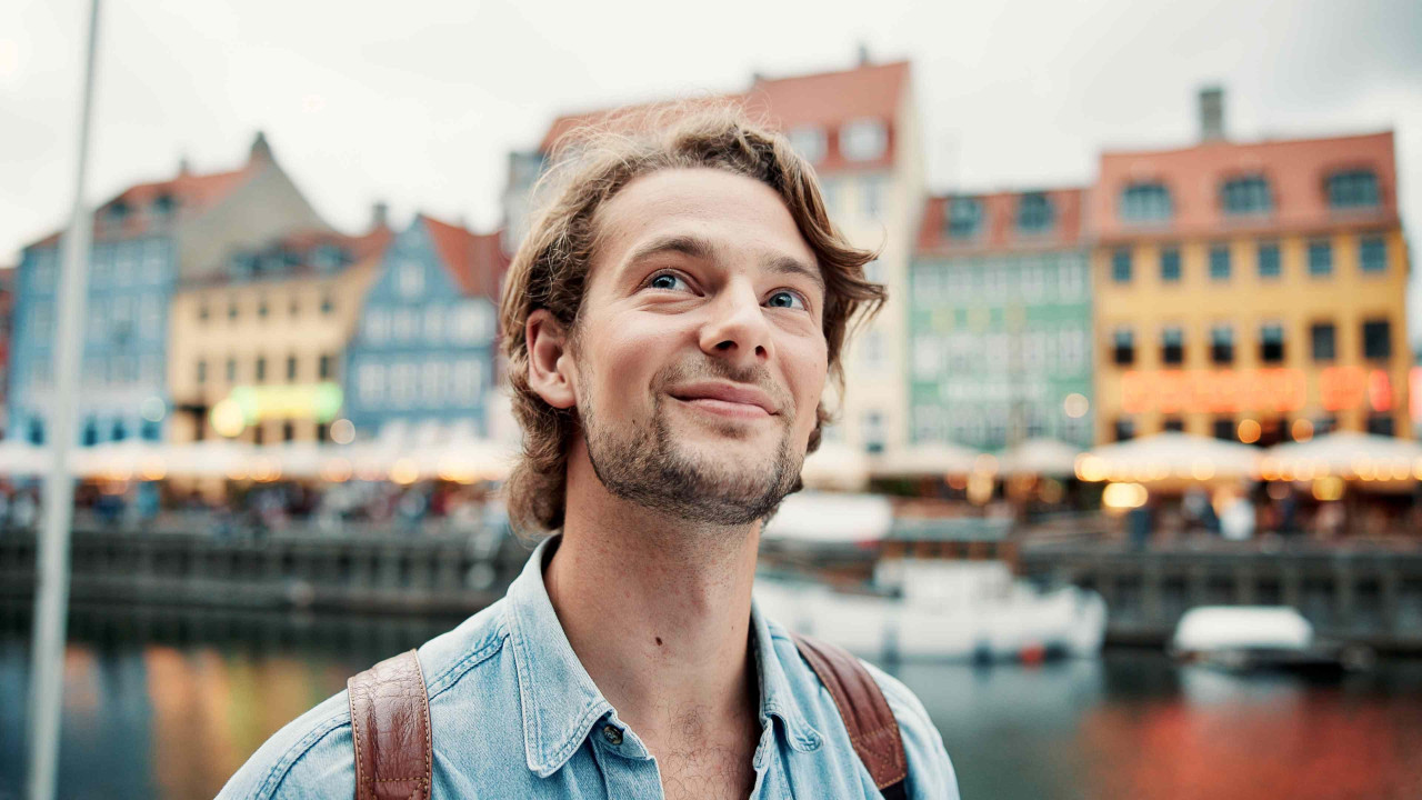 A portrait of a happy wearing a denim shirt and a backpack. He has a happy smile as he looks around. There is a river and colourful buildings in the background. 