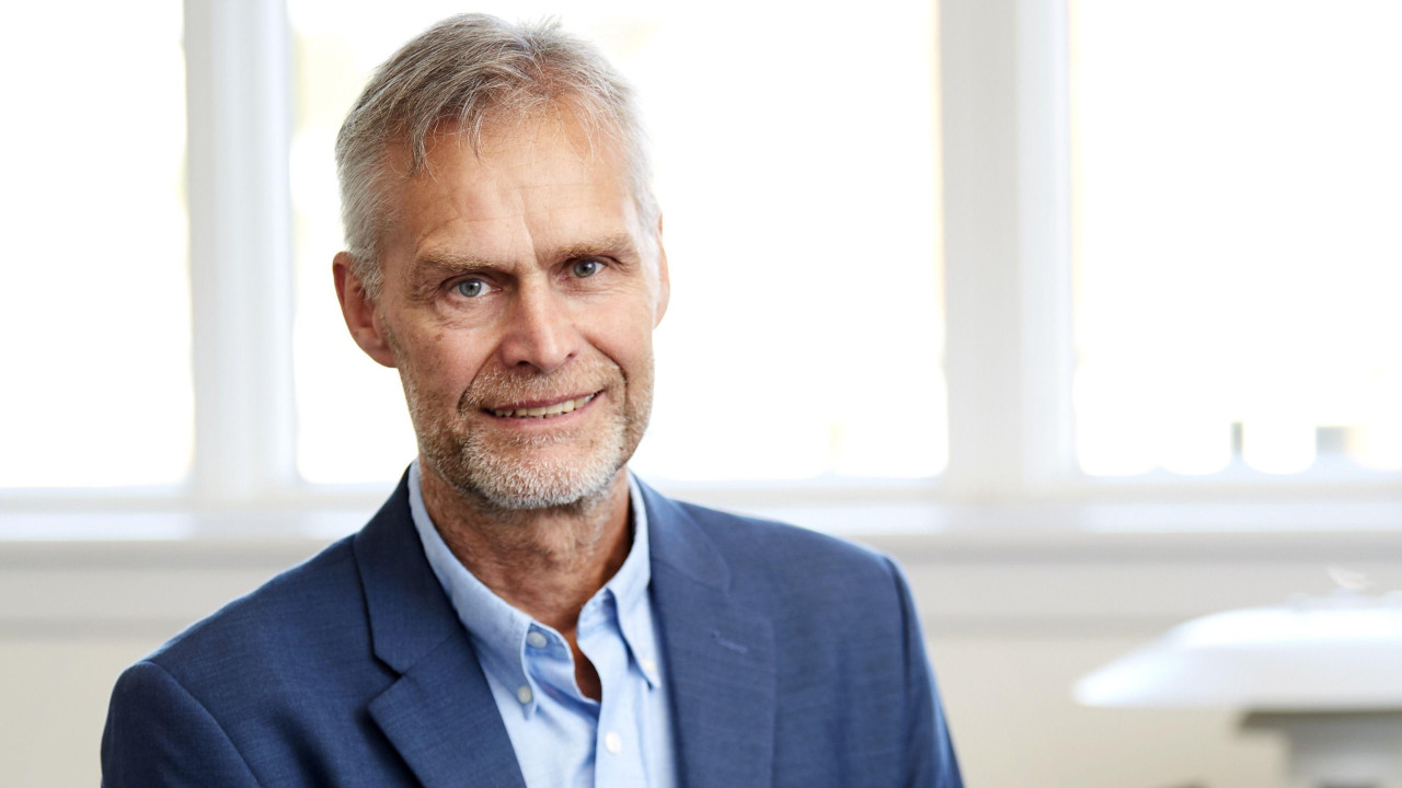 A grey-haired man in a blue shirt and business suit jacket sits smiling at the camera with a clinical setting and big windows blurred in the background