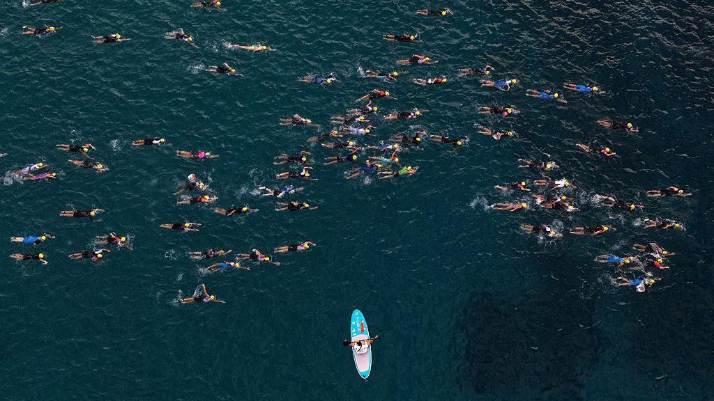 Pogled iz vazduha prikazuje desetine plivača tokom Ironman trke na Havajima, dok ih posmatra osoba u čamcu.