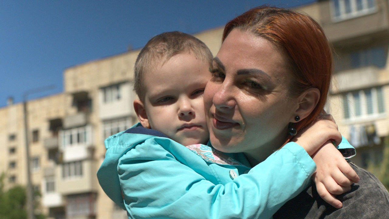 Olena ima ofarbanu crvenu kosu, vezanu pozadi, i nosi sivu majicu. Grli Viktoriju, koja nosi tirkiznu jaknu i ima kratko ošišanu kosu. Nalaze se napolju, a iza njih je stambena zgrada.