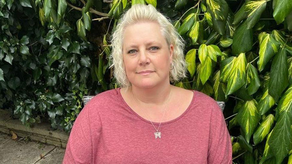 
                    Kler sedi na stolici i gleda pravo u kameru. Ima kratku, plavu, kovrdžavu kosu. Nosi tamnoružičastu majicu kratkih rukava i srebrnu ogrlicu. Napolju je, a u pozadini je ž