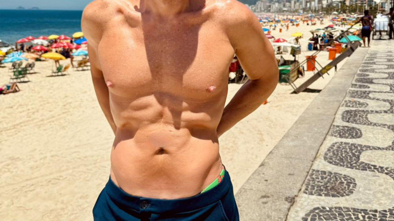 Slikačoveka sa golim, mišićavim torzom na plaži, u plavom šorcu.