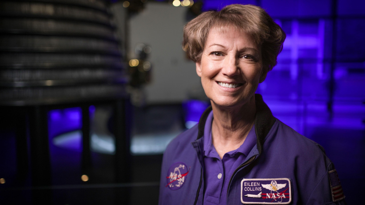 Eileen Collins wearing a blue jacket with a round Nasa patch on one side and a rectangular shuttle patch on the other with her name. She has short brown hair and brown eyes and is smiling as she looks directly to camera. She is in the space gallery at the Science Museum and an out of focus rocket engine can be seen behind her. 