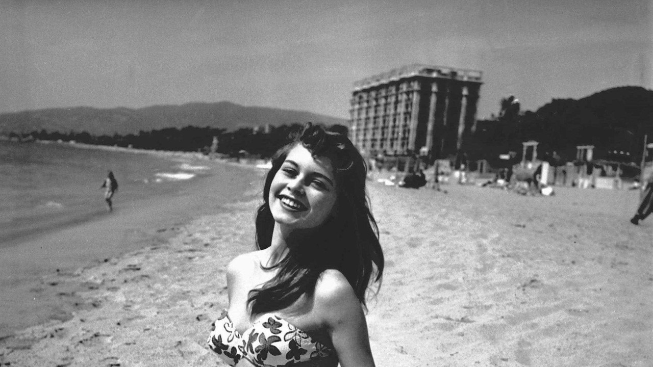 Brižit Bardo na crno-beloj fotografiji, sa dugom tamnom kosom, u cvetnom bikiniju, sedi na pesku i smeši se fotografima na Filmskom festivalu u Kanu 1953. godine.