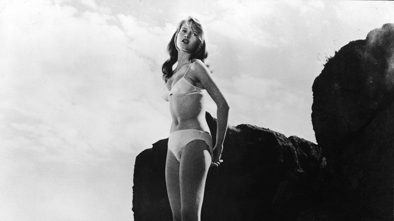 Francuska glumica Brižit Bardo nosi beli bikini i stoji na steni pored mora na kadru iz filma Devojka u bikiniju, u režiji Vilija Rozihera iz 1958. godine.