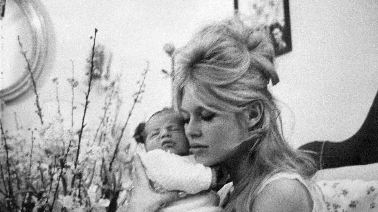 Brižit, obučena u noćnu haljinu, sa plavom kosom stilizovanom u visoku punđu, nežno drži sina blizu lica.