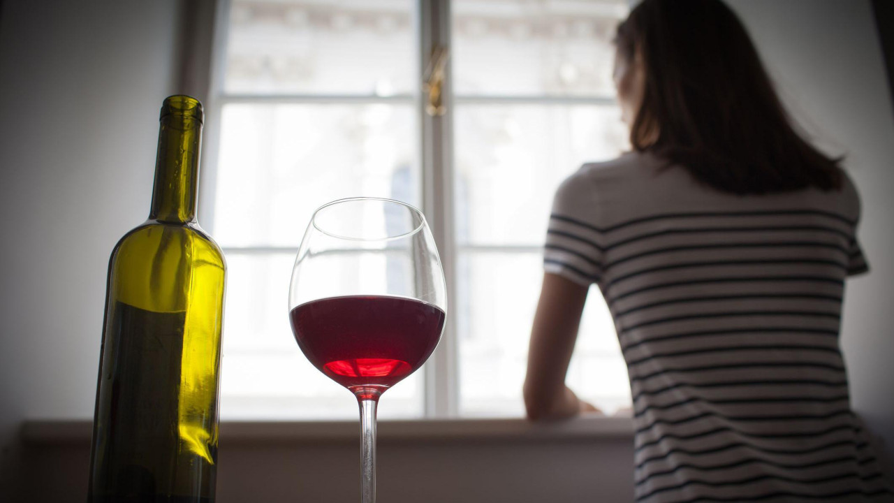 Prazna boca vina uz polupunu čašu, dok u pozadini žena gleda kroz prozor