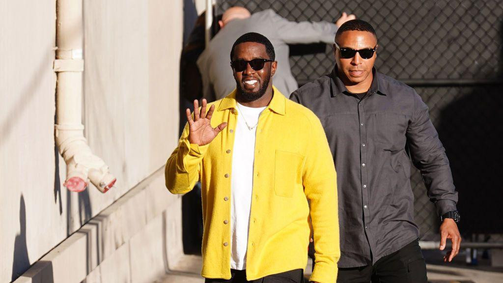 Šon „Didi“ Kombs, obučen u žutu majicu i tamne pantalone, sa naočarima za sunce, maše kamerama i osmehuje se pre nego što je uhapšen. Muškarac obučen u crno - takođe sa naočarima za sunce - hoda pored njega.