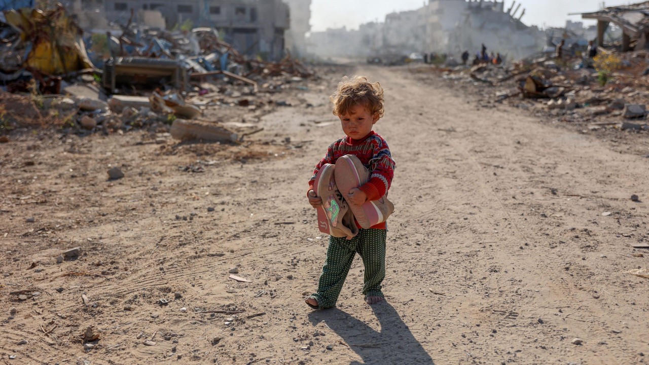 Palestinsko dete sa igračkama u rukama na ulici, okruženo ruševinama, u šarenom džemperu i zelenim pantalonicama 
