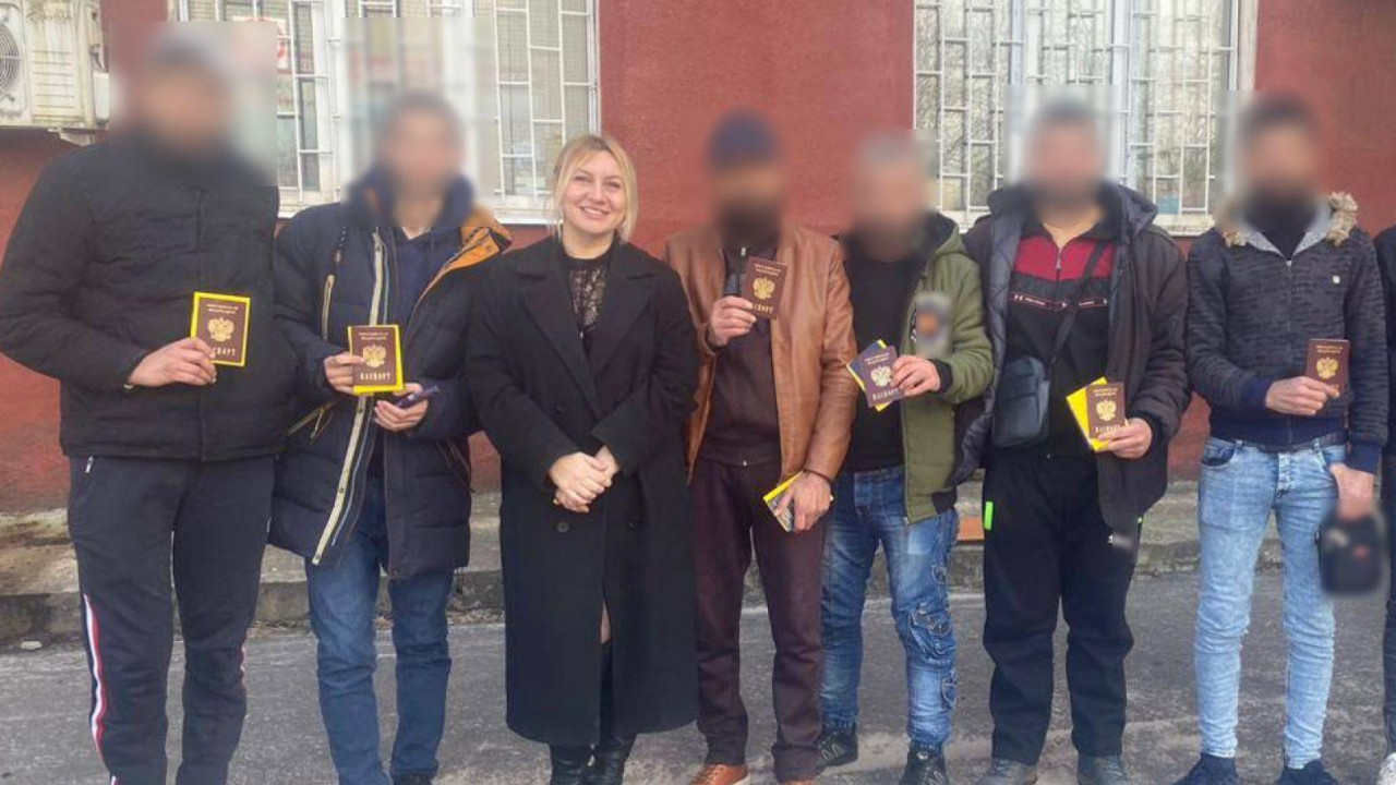 Polina Azarnik stoji nasmejana u dugom crnom kaputu i ​​elegantnim crnim čizmama u društvu muškaraca u farmerkama, cipelama i jaknama. Njihova lica su zamagljena kako bi se sakrio njihov identitet. Drže ruske putne isprave.