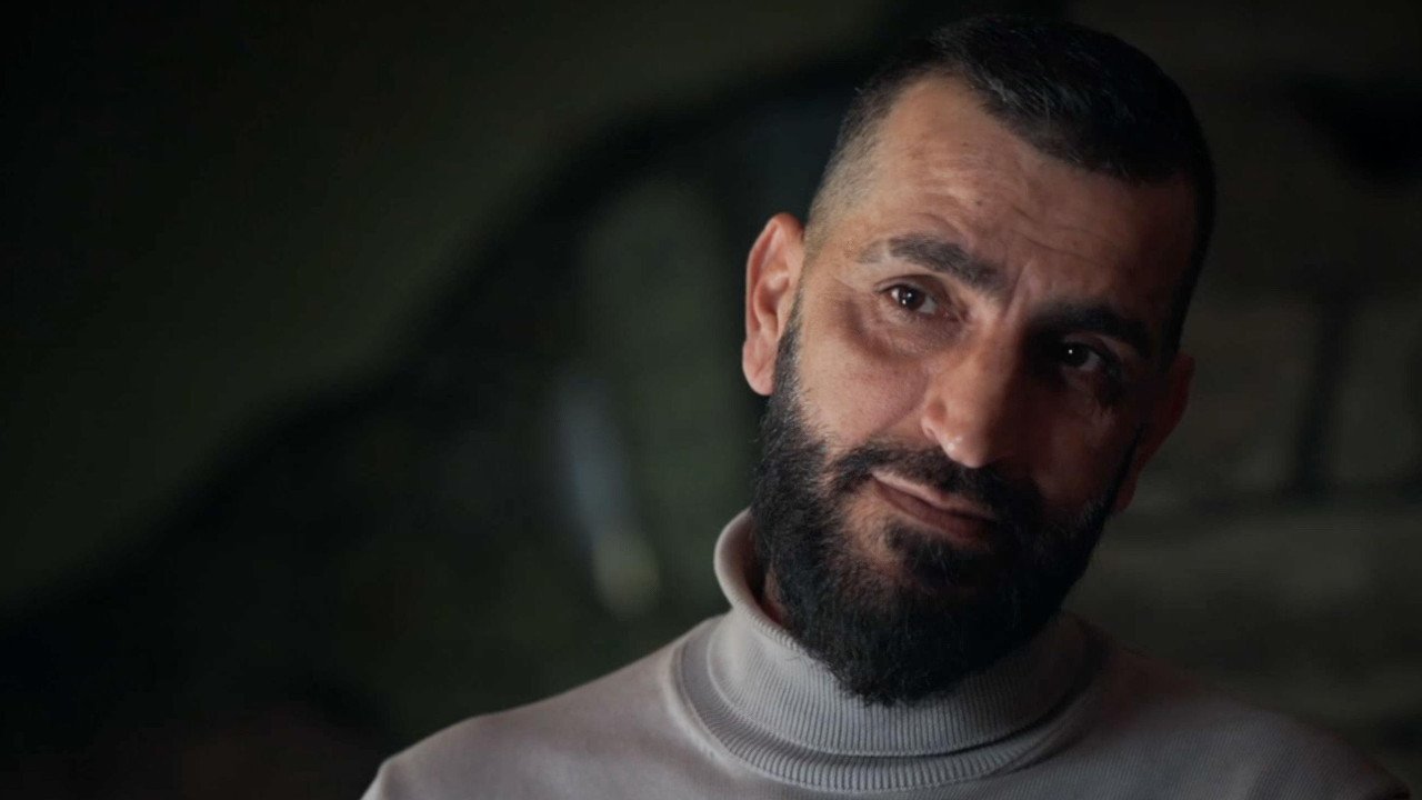 Habib u beloj majici sa rol kragnom gleda u kameru. Ima tamnu bradu i brkove.
