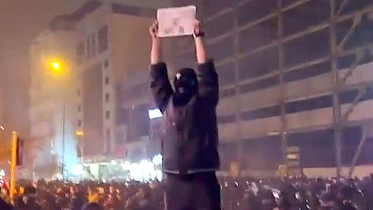 
                    A masked protester stands on a platform above crowds at night in Kaj Square, in north-west Tehran. on 9 January 2026. They are holding a black and white photo above his h