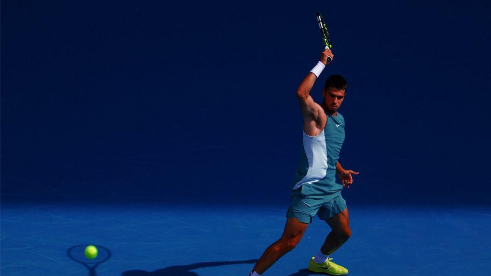 Teniser Karlos Alkaraz udara lopticu tokom meča četvrtog kola protiv Britanca Džeka Drejpera na Australijan openu 2025. godine. Nosi tirkiznu opremu i neonsko žute patike na jarko plavom terenu, a njegova senka izgleda kao da udara lopticu.