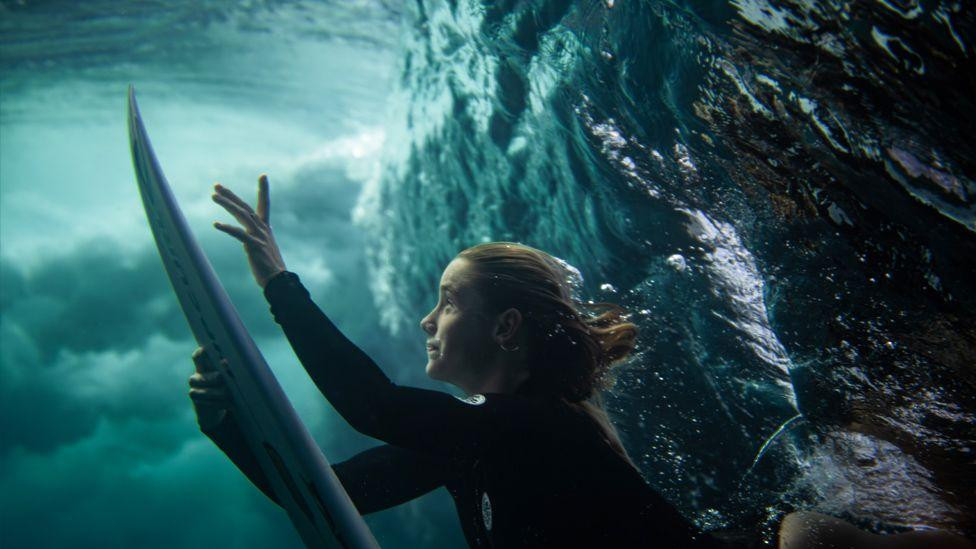 Podvodni pogled na surferku koja gura dasku za surfovanje nadole dok roni ispod talasa. Voda je bistra sa vrtložnim mehurićima, a surferka nosi crno odelo za ronjenje.