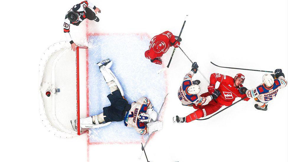 Pogled odozgo na hokejašku utakmicu. Golman leži na ledu dok se igrači u crveno-plavim dresovima bore za pak oko gola.