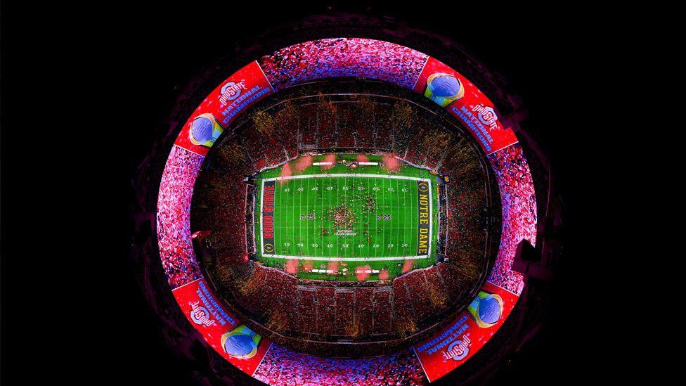 Pogled iz perspektive ribljeg oka na jarko osvetljeni fudbalski stadion pun navijača. Zeleni teren je u centru, okružen jarko crvenim i plavim transparentima sa nazivima timova i grafikom prvenstva. 