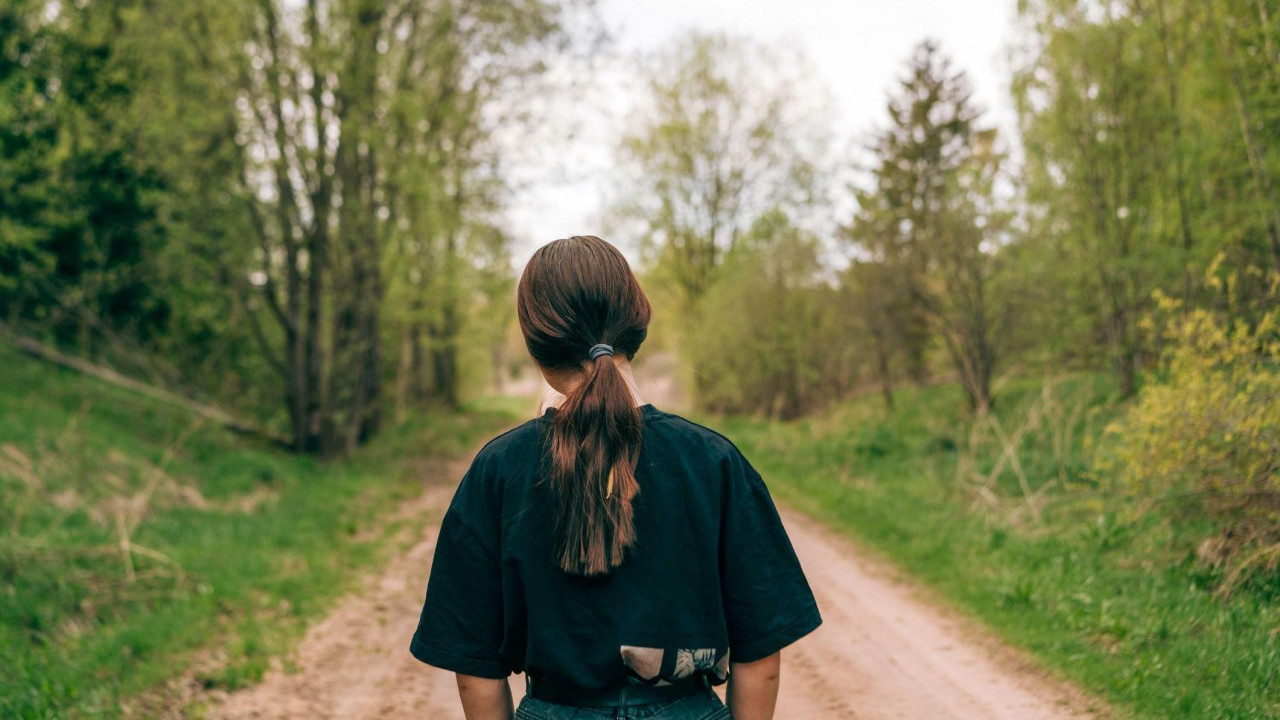 devojka sa skupljenom kosom okrenuta leđima stoji na putu kroz šumu 