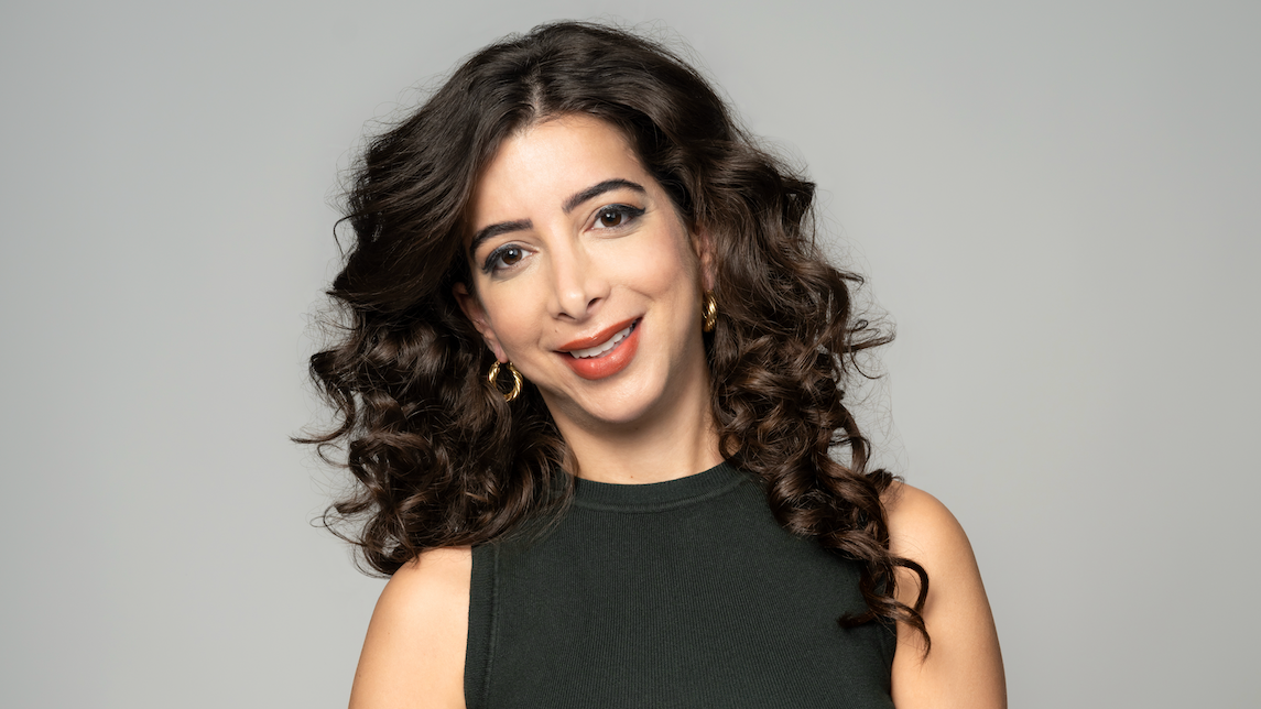 Žena duge tamne, kovrdžave kose, sa zlatnim okruglim minđušama na ušima i crvenim karminom, nasmejana gleda u kameru, glave blago nagnute na levo, u tamnoj majici bez rukava