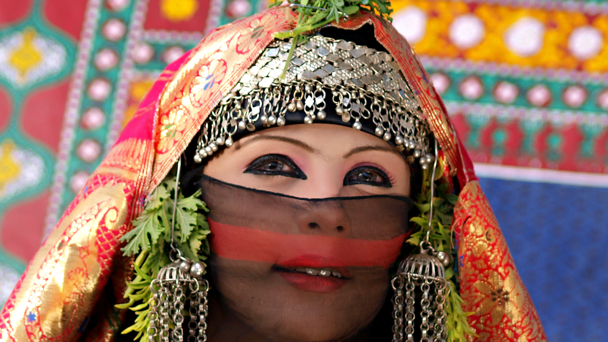 Beduinka obučena u šarenu, mahom crvenu garderobu, prekrivene glave i lica šarenom providnom tkaninom i metalnim ukrasima sa obe strane, obojenih očiju kajalom