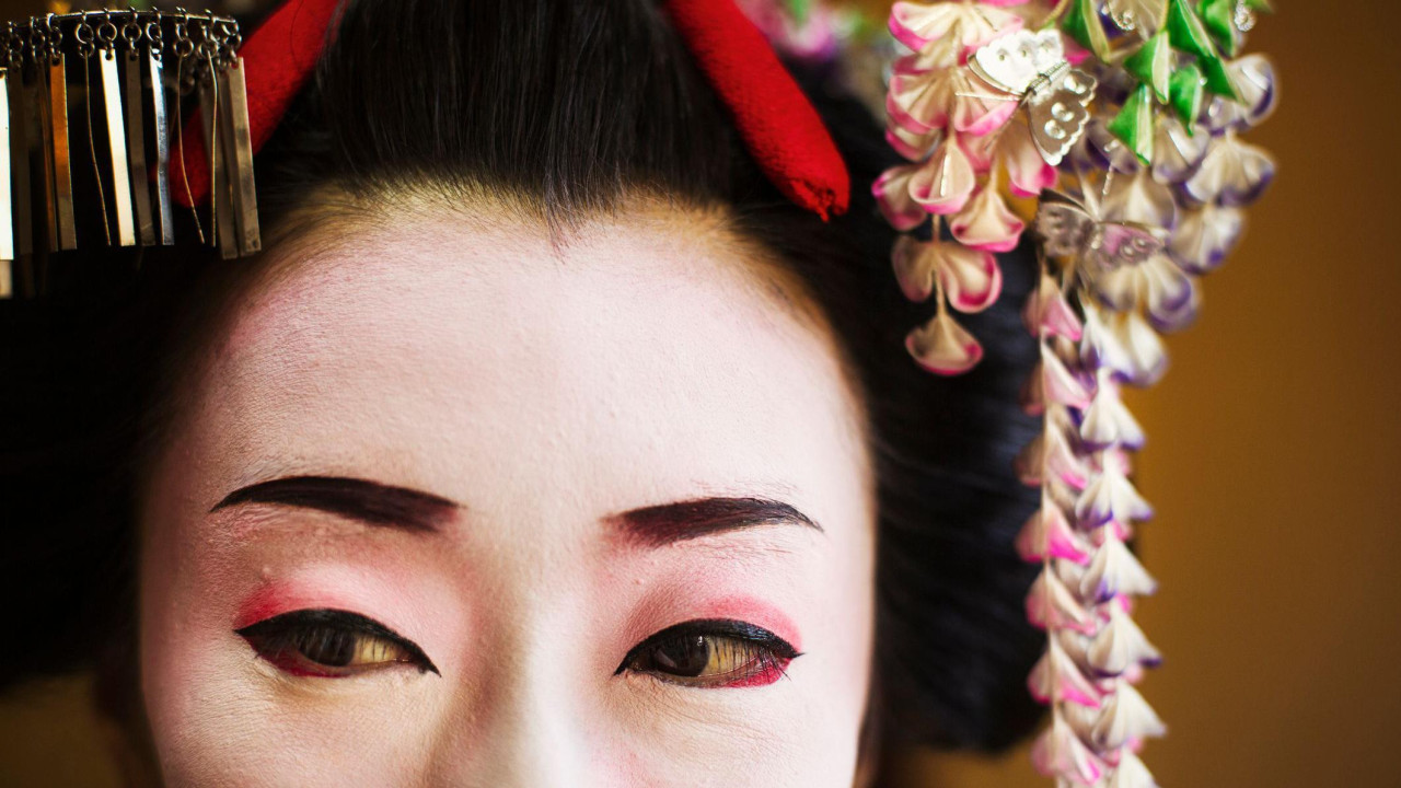 Fotografija japanske gejše - prikazan je samo gornji deo lica - oči, našminkane crnom i crvenom bojom, sa ukrasima oko lica i na glavi u belim, roze i ljubičastim tonovima