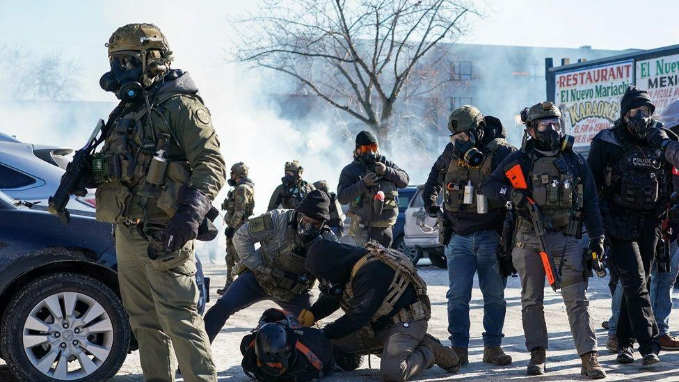 Čovek leži na ulici sa snegom, a dvojica agenata kleče pore njega i vezuju mu ruke