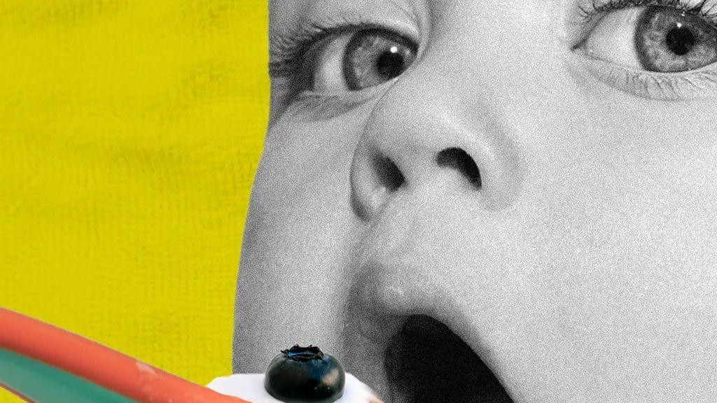 Beba otvara usta i gleda u kameru dok je hrane kašičicom jogurta, a preko nje je borovnica. Pozadina je žuta, a kašičica narandžasta i zelena