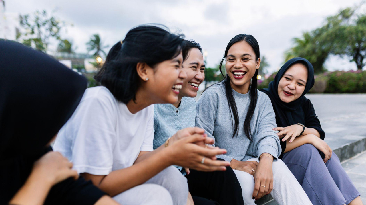 Pet devojaka sede na trotoaru i smeju se 