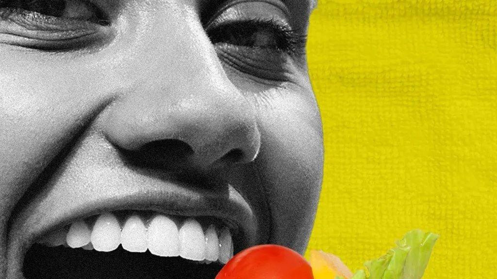 Žena gleda u kameru dok ustima prinosi viljušku sa paradajzom, paprikom i zelenom salatom