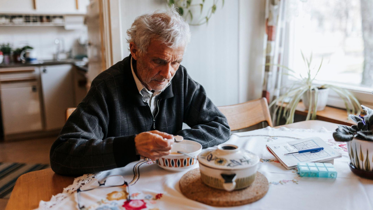 Stariji čovek sa sedom kosom sedi za stolom u kući i jede žitarice iz činije