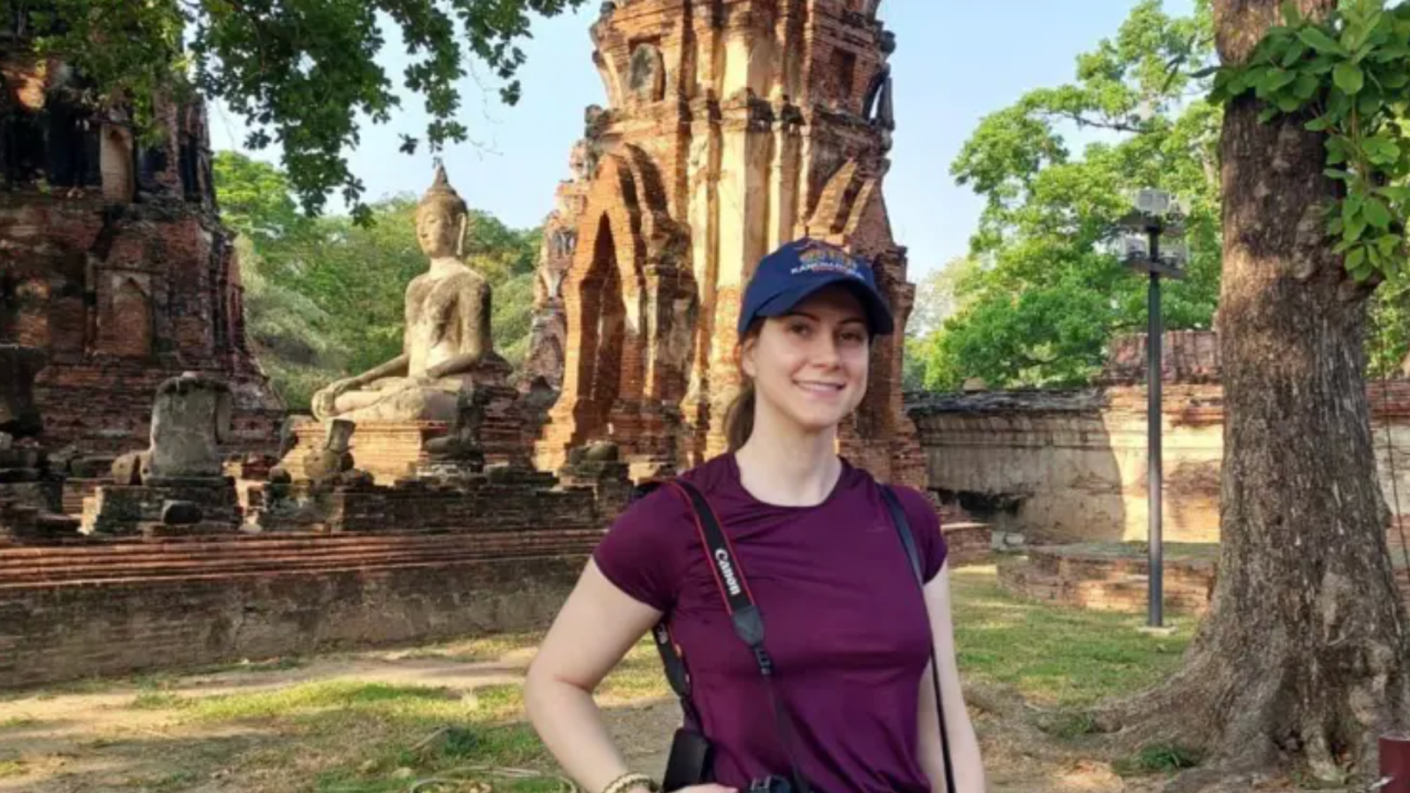 Devojka sa bejzbol kapom u ljubičastoj majici i fotoaparatom oko vrata ispred statue Bude na Tajlandu